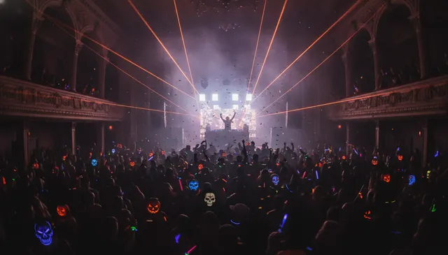 Verkleidete Menge mit leuchtenden Kürbismasken bei einem Halloween-Rave unter orangefarbenen Lasern: Erstelle dein eigenes BOO-Seattle-Musikquiz-Paket (Hitster-Style) mit Hitify
